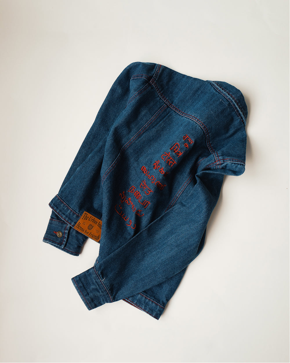 Khadi denim jacket in Medium Blue folded at an angle, sleeve and back embroidery in red thread with the leather cuff patch