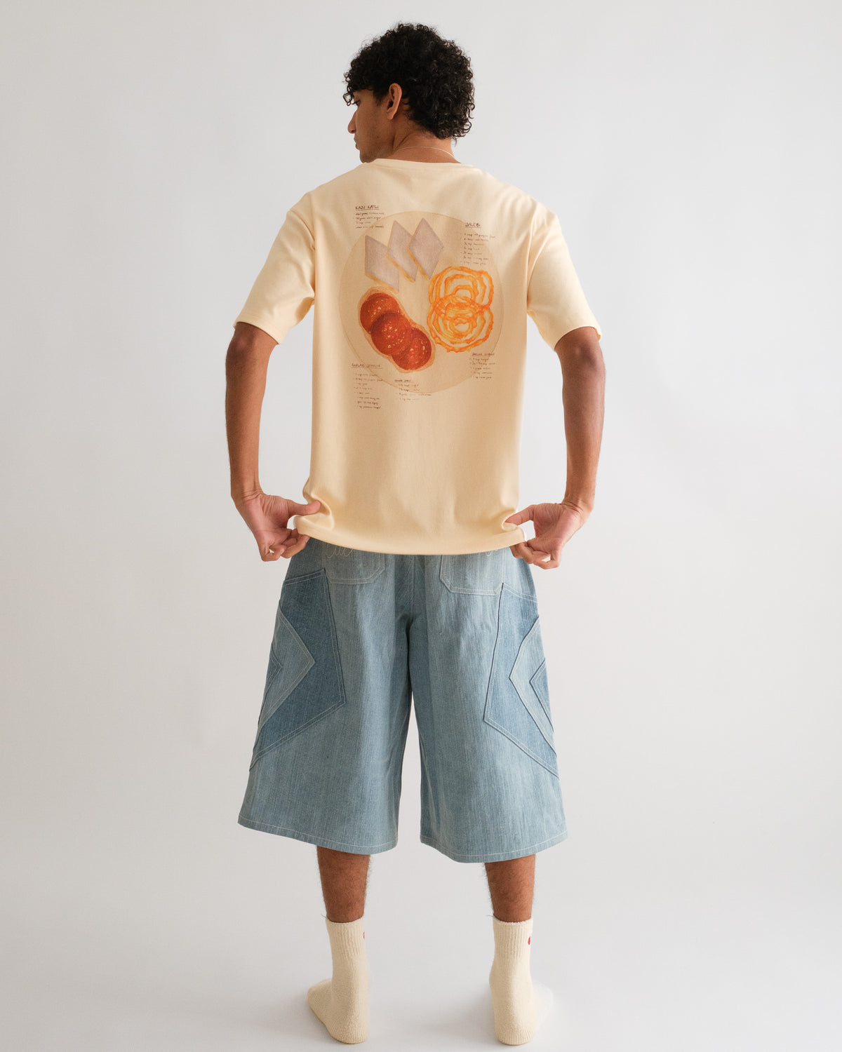 Man from behind pulling the hem of the Recipe Tee to show the back print, with the Khadi Katli Jorts