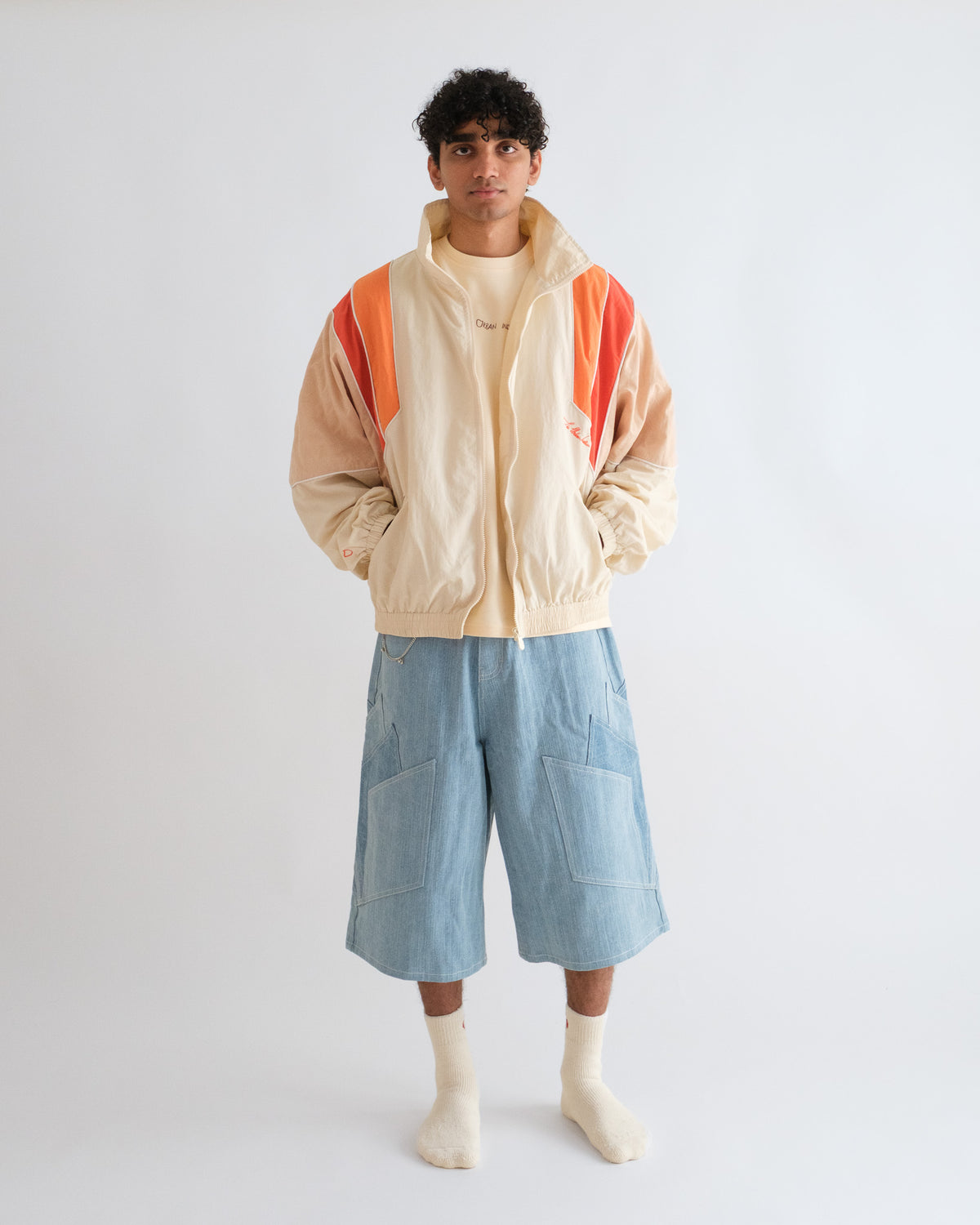 Man wearing the Sweets Track Jacket in Saffron Orange open over a cream tee, with the Khadi Katli Jorts #Color_Saffron Orange